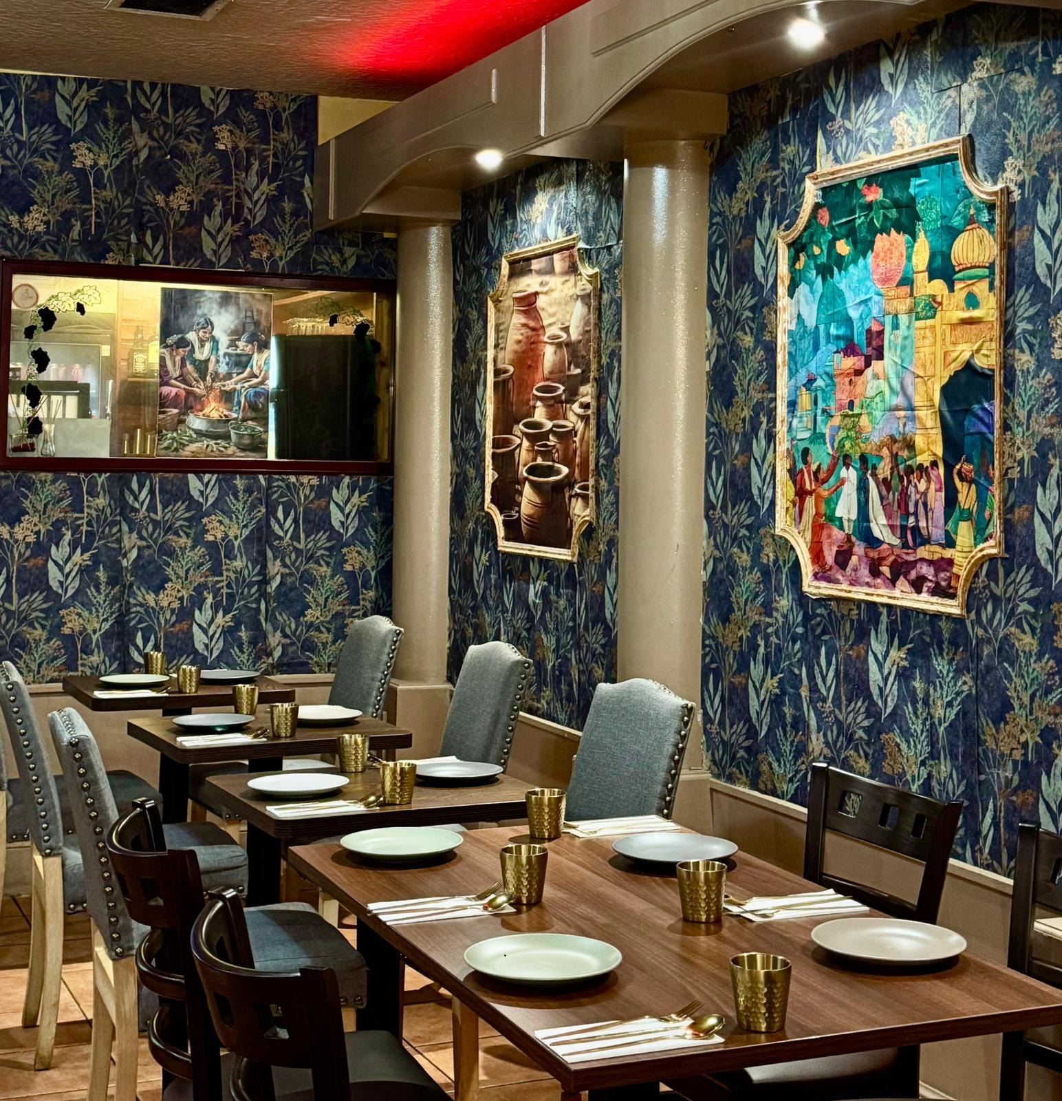Dining room at Gumnam: tables set with gold tumblers beneath framed Indian artwork on navy botanical wallpaper